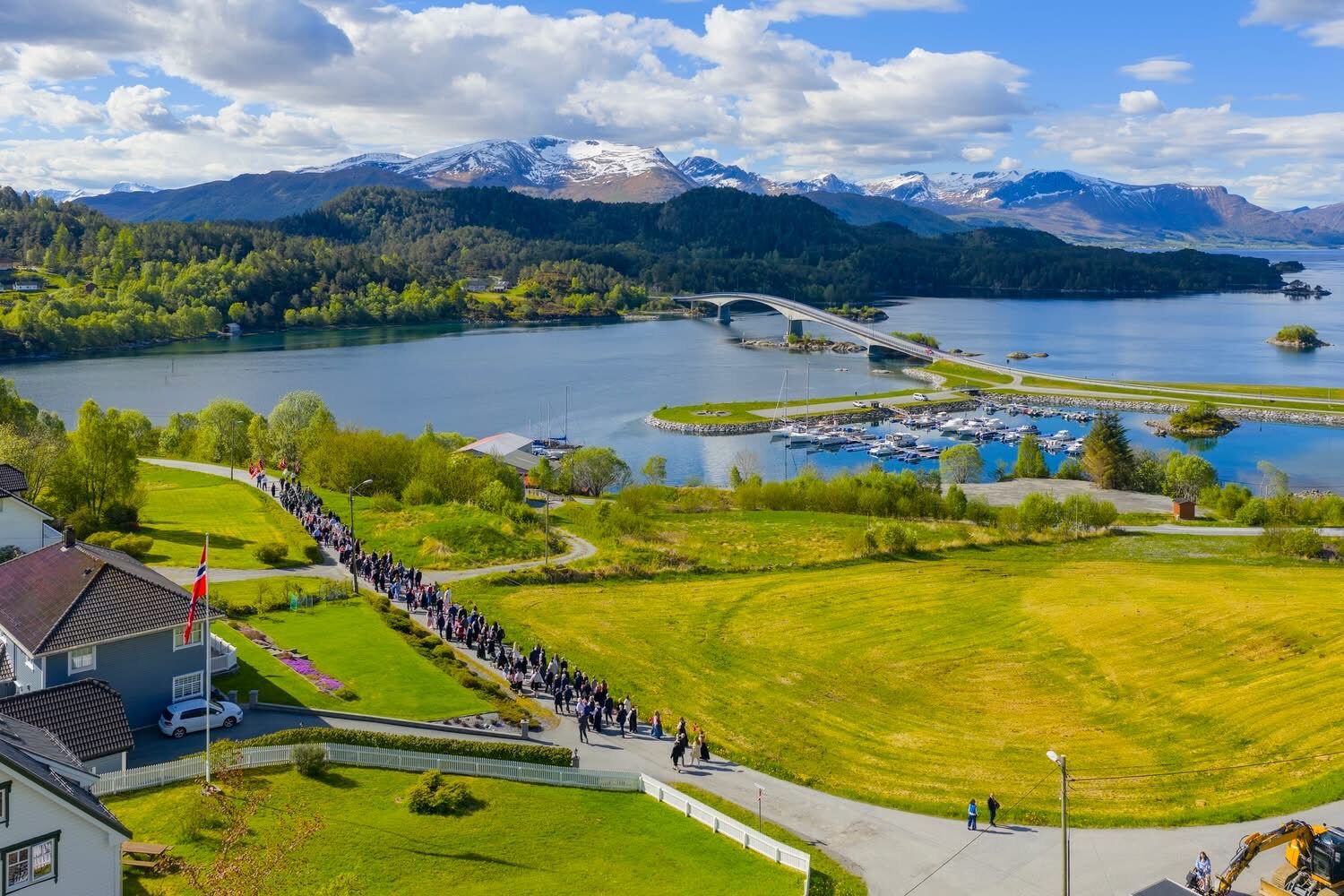 17. mai-tog med bunadskledde bygdefolk på veg mot grendahuset, med Eiksundbrua og fjella i bakgrunnen